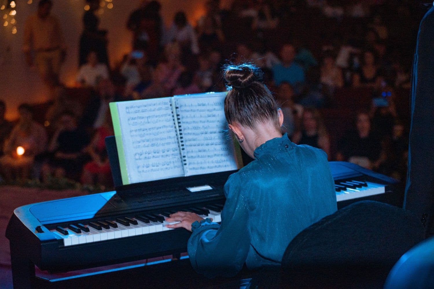 A young performer in a teal dress at a digital piano before a candlelit audience