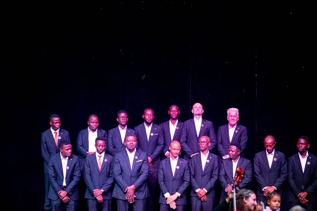 Men of the ensemble standing formally on a darkened stage