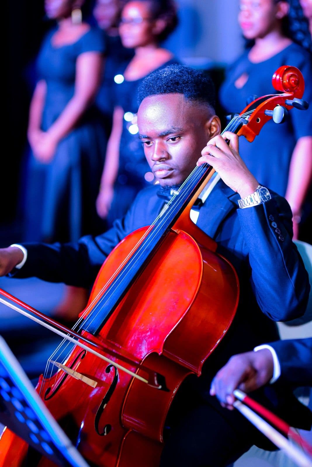 A cellist in a tuxedo playing on stage with the choir behind him