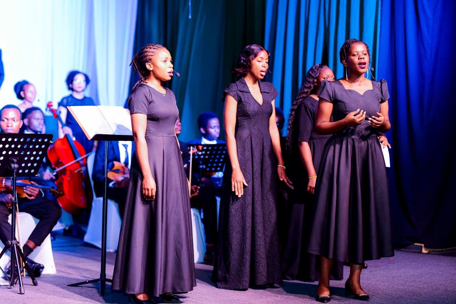 Three female vocalists in black gowns singing with orchestra behind them