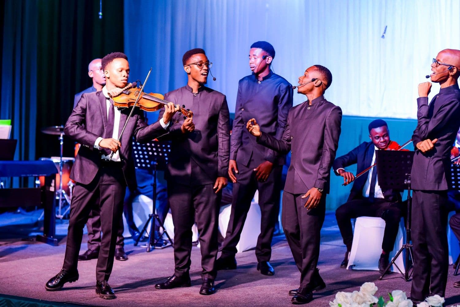 A violinist and male singers in matching formal wear under blue lighting