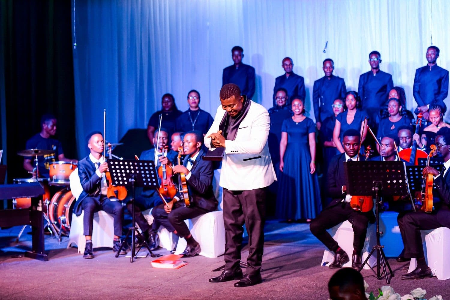 A conductor in a white tuxedo jacket bowing with orchestra and choir behind him
