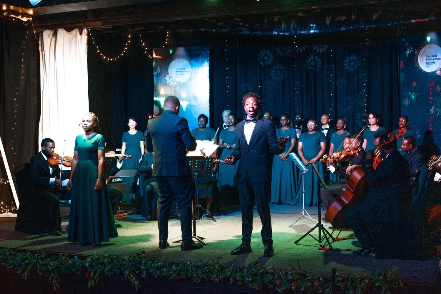 Male and female soloists with choir, strings, and conductor under holiday lights